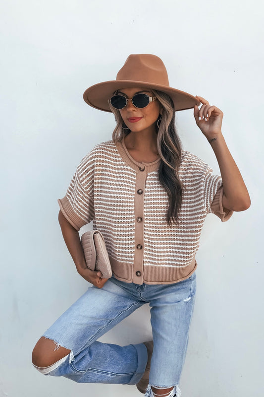 Woman in tan striped button down sweater, hat, and sunglasses poses with clutch and ripped jeans against a white wall.