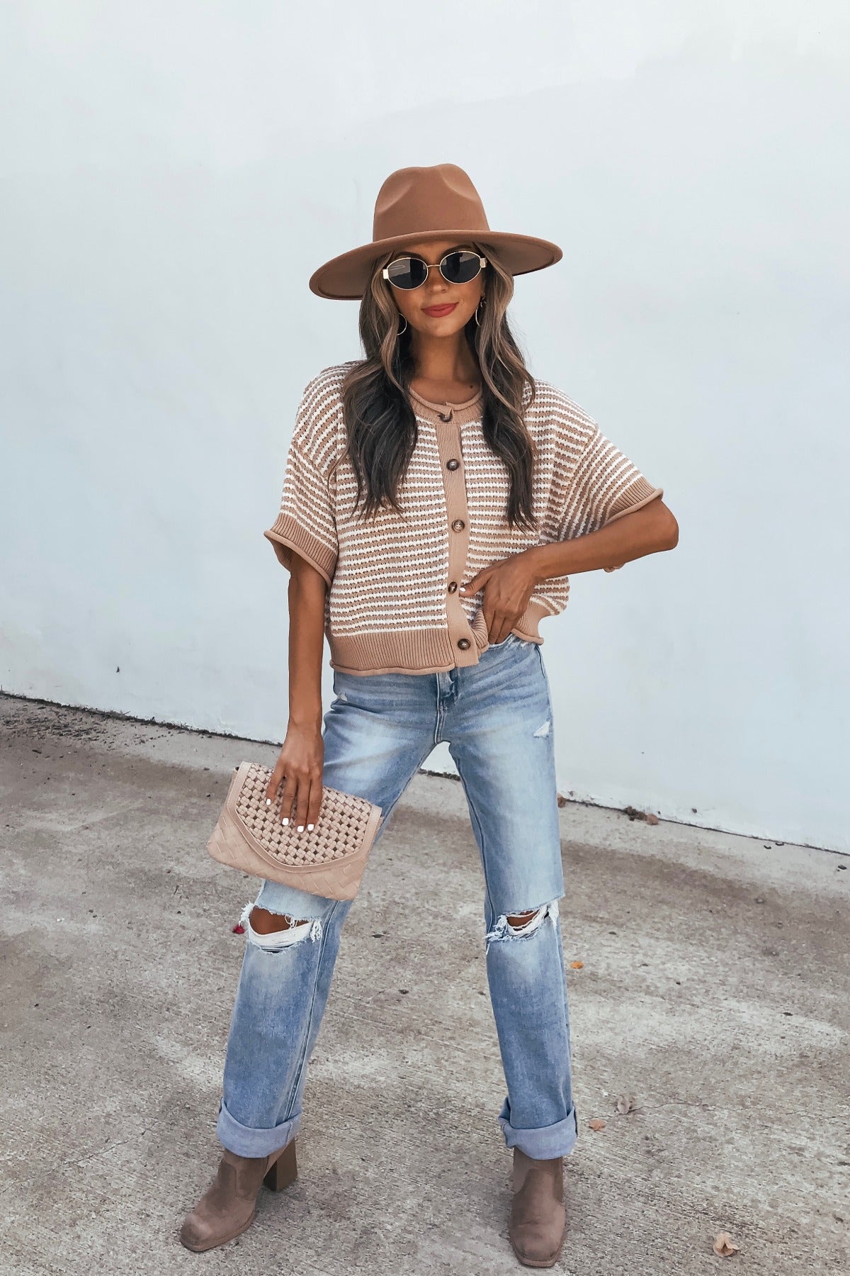 Woman in a tan hat, Tan Striped Button Down Sweater, ripped jeans, and brown boots holds a beige clutch by a plain wall in a crochet cardigan.