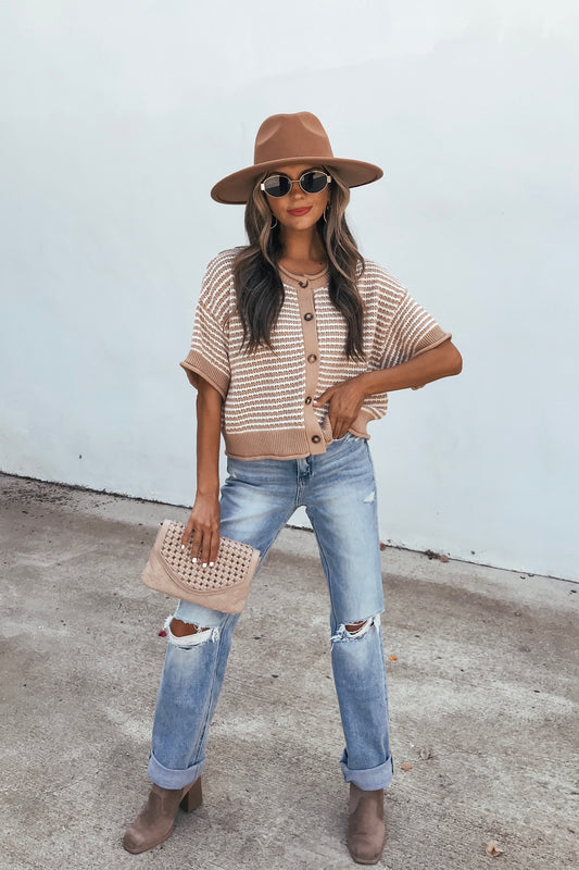 Woman in a tan hat, Tan Striped Button Down Sweater, ripped jeans, and brown boots holds a beige clutch by a plain wall in a crochet cardigan.