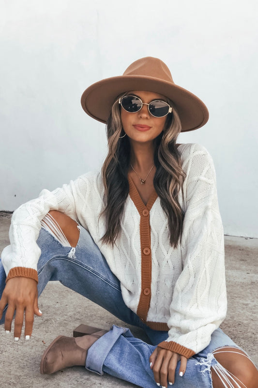 Woman in a cream contrast trim cardigan, wide-brim hat, sunglasses, ripped jeans, and tan shoes sits on pavement by a white wall.