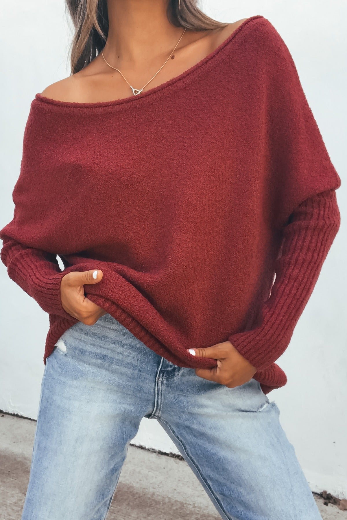 Woman wearing an Off The Shoulder Ribbed Sweater in wine and light blue jeans, standing outside against a white wall.