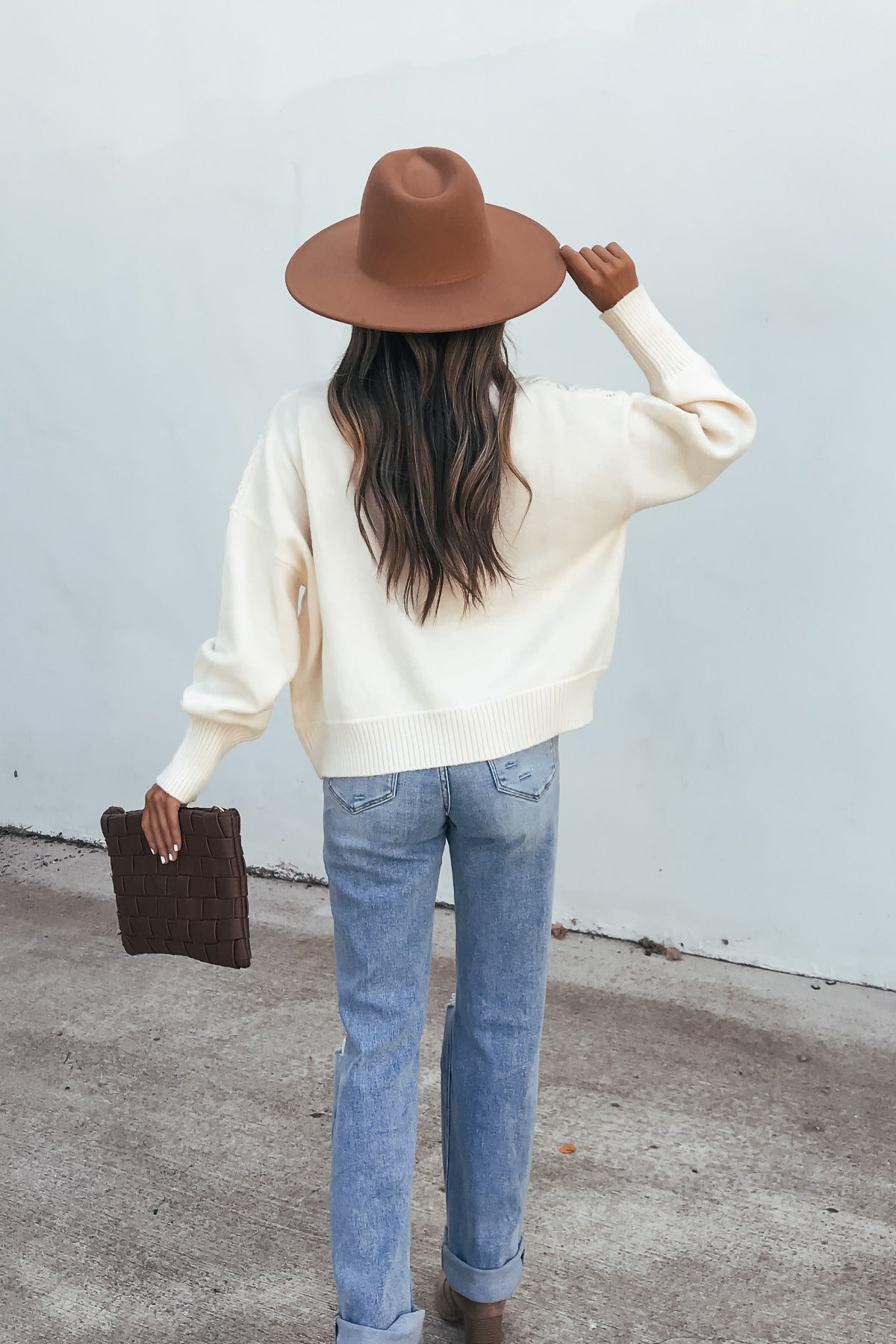 Woman in a Cream Mixed Knit Sweater, brown hat, jeans, and holding a brown clutch stands on concrete facing away.