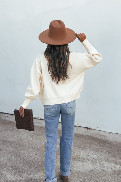 Woman in a Cream Mixed Knit Sweater, brown hat, jeans, and holding a brown clutch stands on concrete facing away.