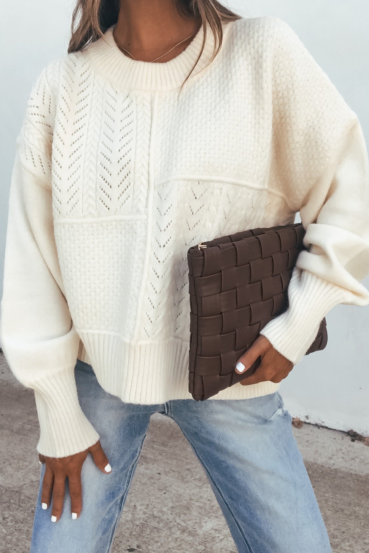 Woman in a Cream Mixed Knit Sweater and light jeans holds a brown clutch bag while standing outdoors.