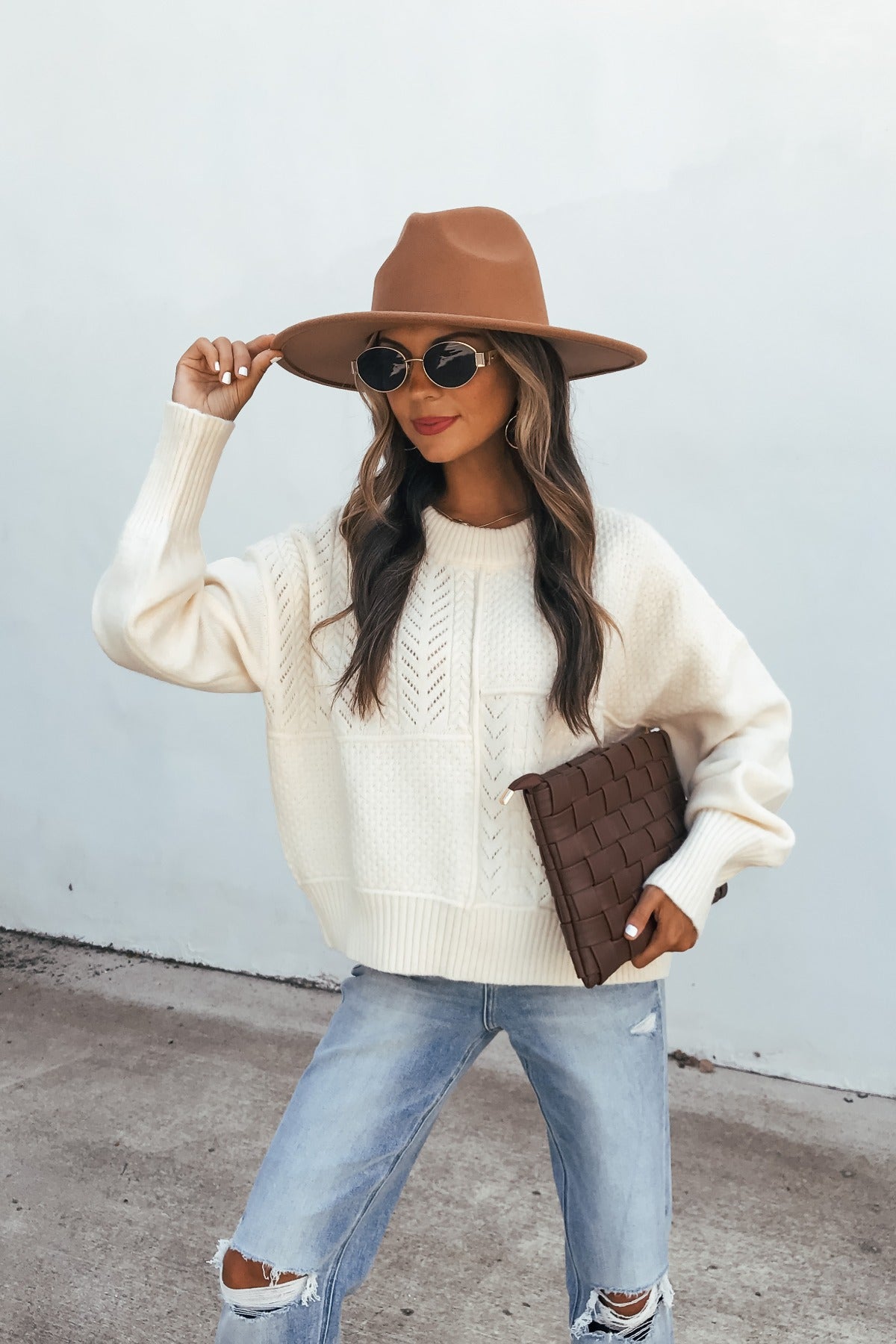 Woman in a Cream Mixed Knit Sweater, brown hat, and ripped jeans holds a woven clutch against a light background.