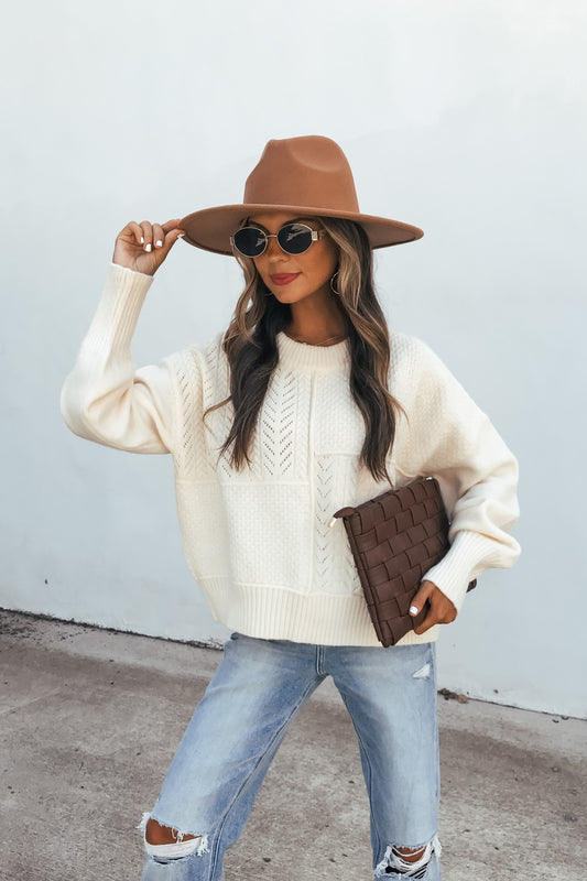 Woman in a Cream Mixed Knit Sweater, brown hat, and ripped jeans holds a woven clutch against a light background.
