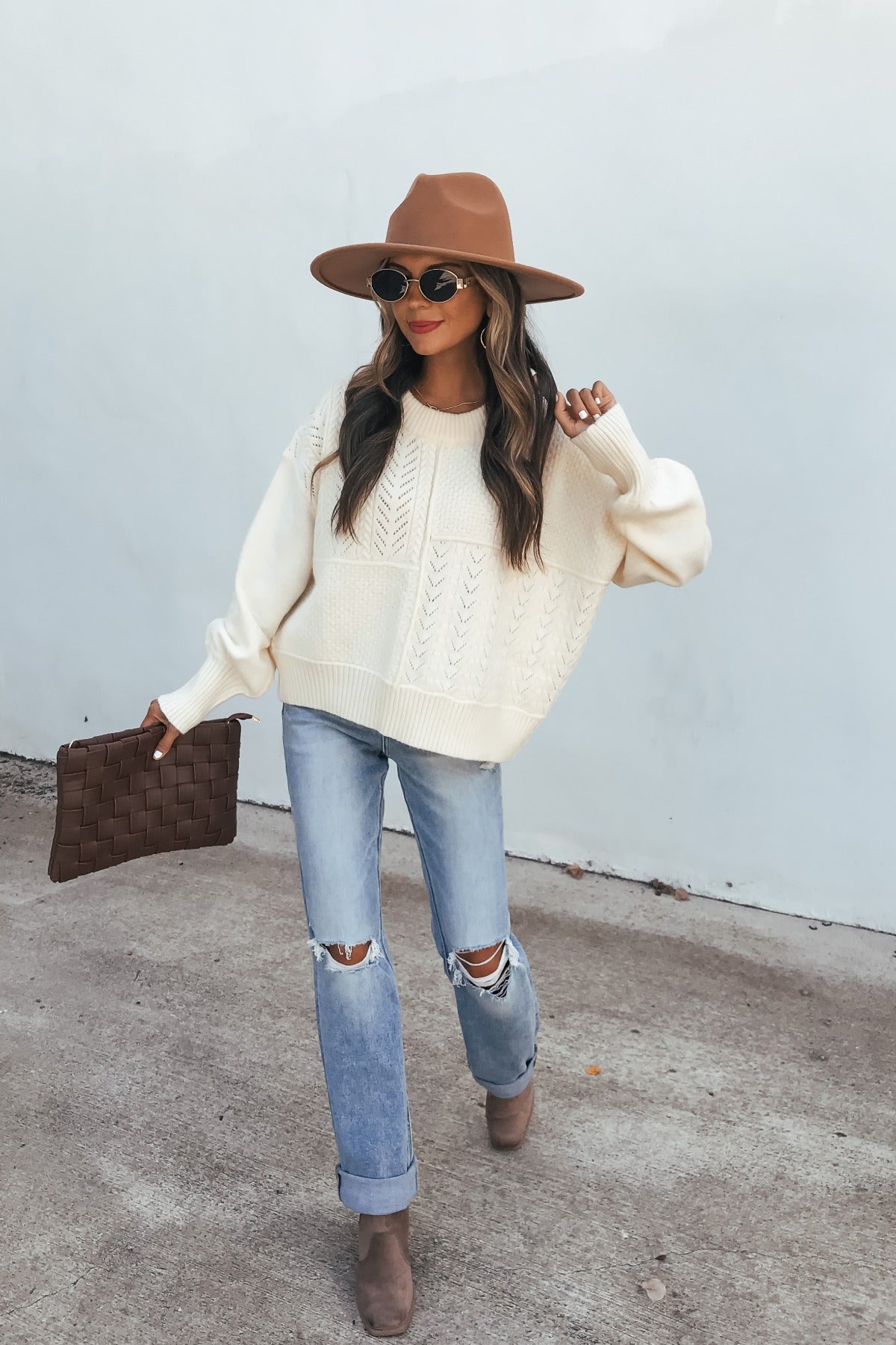 Woman in a Cream Mixed Knit Sweater with ripped jeans, boots, and a brown clutch walks by a light wall.