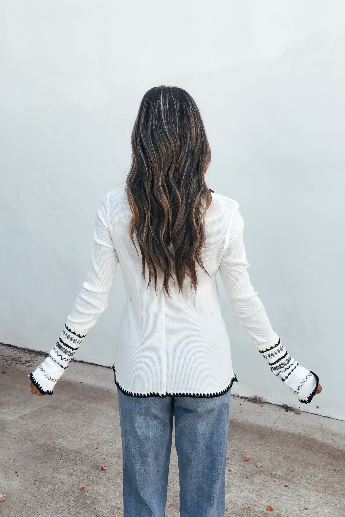 Woman with long wavy hair wears a white Free People Aspen Thermal Top and blue jeans, standing by a plain light wall.