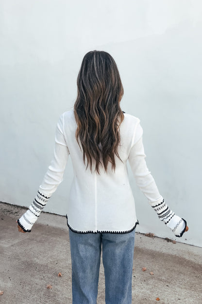 Woman with long wavy hair wears a white Free People Aspen Thermal Top and blue jeans, standing by a plain light wall.