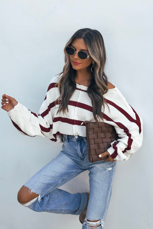 Woman in sunglasses models the Burgundy Striped Dolman Sleeve Sweater with ripped jeans and a brown woven clutch against a white background.