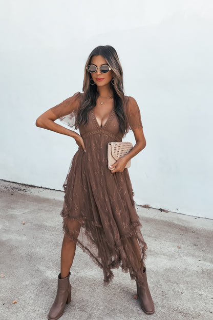 Woman in an Embellished Lace Midi Dress - Brown, with boots and clutch, sunglasses on, stands by a plain light wall.