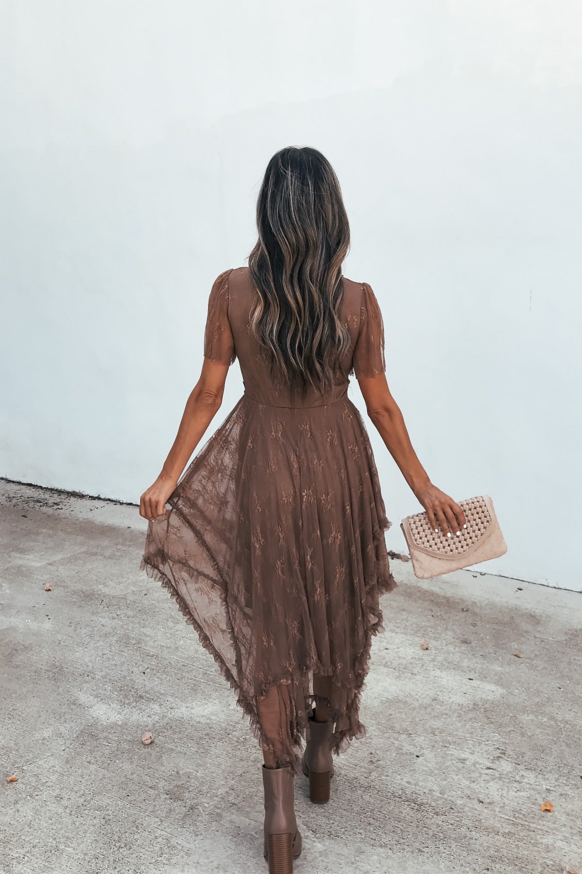 Woman in the Embellished Lace Midi Dress - Brown holds a beige clutch, standing on concrete facing away from the camera.