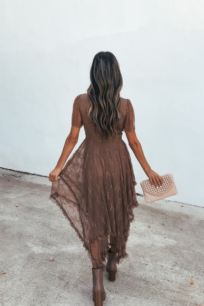 Woman in the Embellished Lace Midi Dress - Brown holds a beige clutch, standing on concrete facing away from the camera.