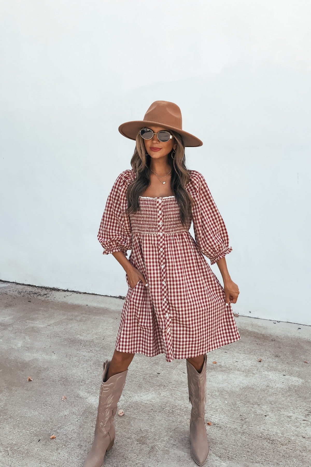 Woman in a brown hat and sunglasses wears the Gingham Babydoll Mini Dress - Burgundy, posing outdoors against a plain white wall.