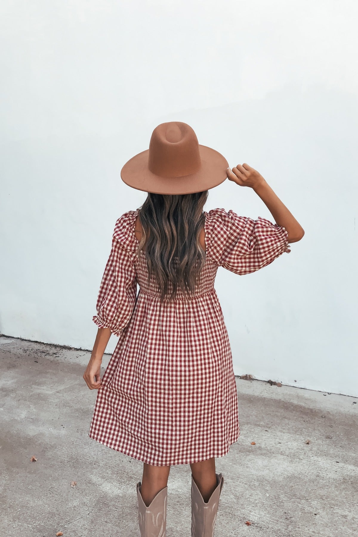 Woman in a Gingham Babydoll Mini Dress - Burgundy, hat, and boots stands on concrete facing a white wall, holding her hat.