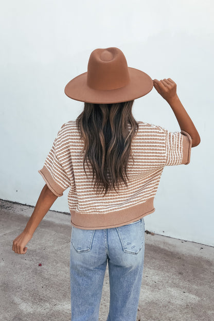 Woman with long wavy hair wears a Tan Striped Button Down Sweater and jeans, brown hat on, standing outdoors facing away.
