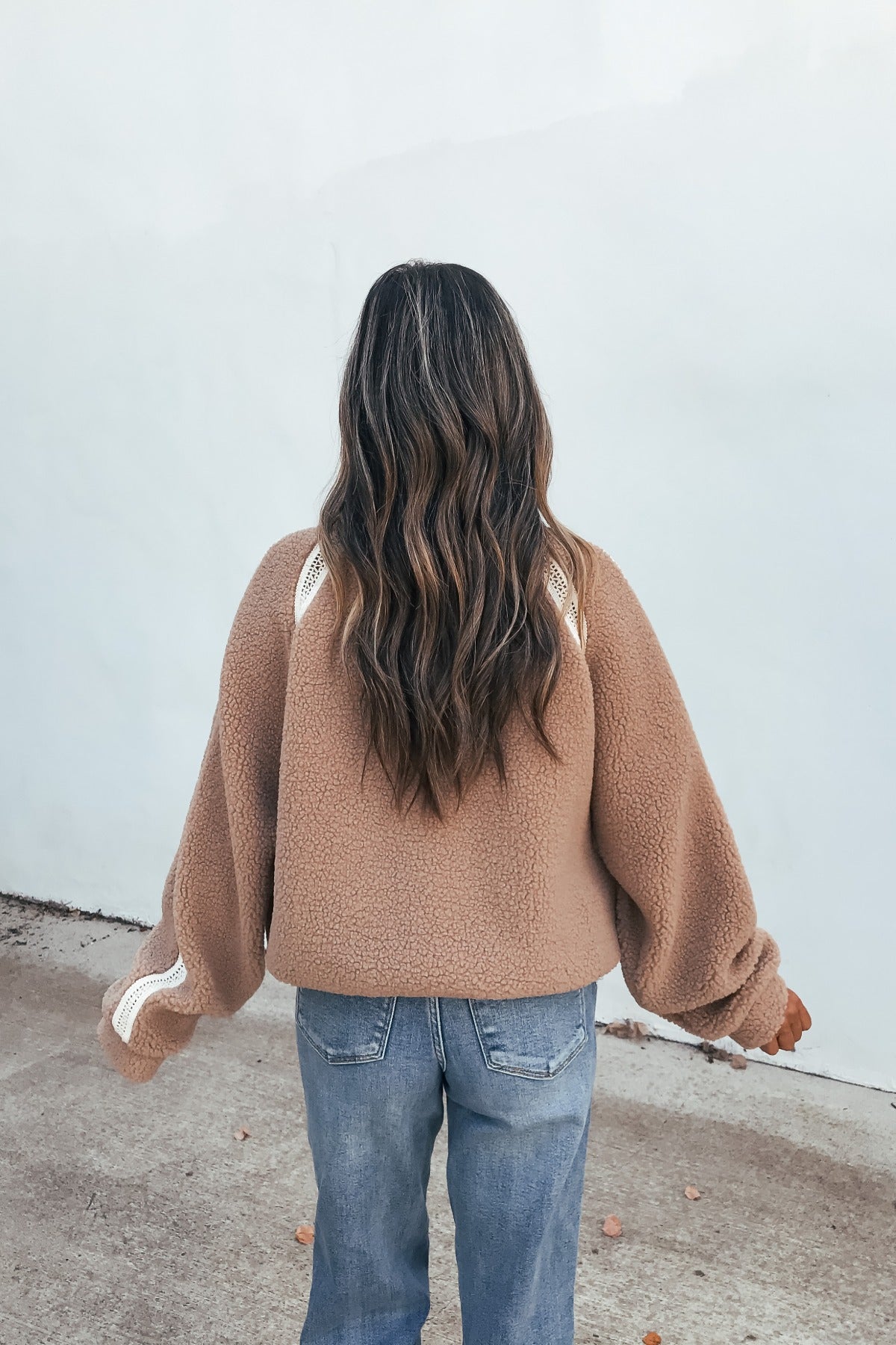 A person with long wavy hair wears the Contrast Quarter Zip Jacket - Sand, paired with blue jeans, facing a white wall.