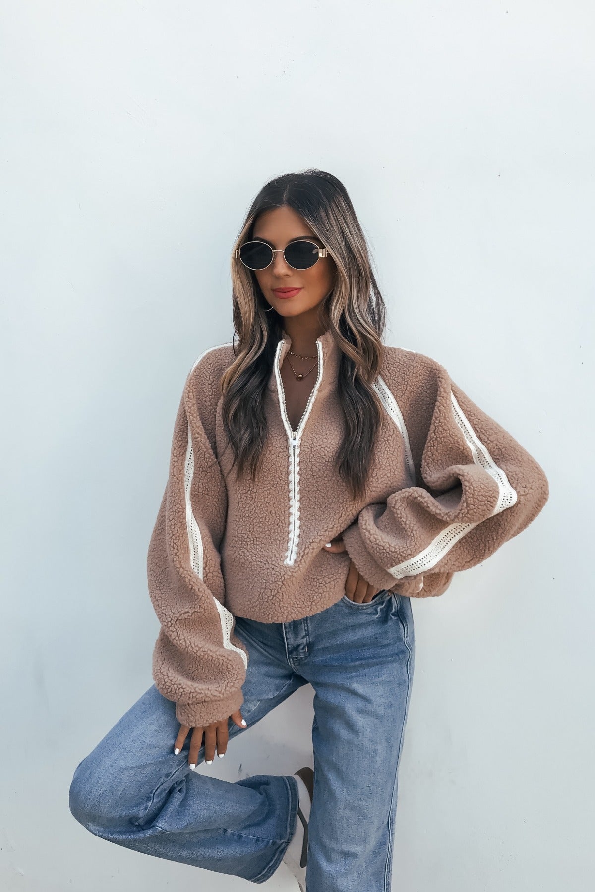 Woman in a Contrast Quarter Zip Jacket - Sand and jeans poses against a white wall, wearing sunglasses with her hair down.