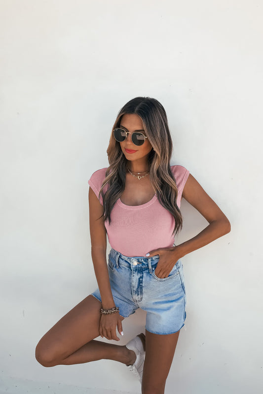 Woman in sunglasses and denim shorts wears a slim Dusty Mauve Scoop Neck Ribbed Top, standing by a white wall with one hand in her pocket.
