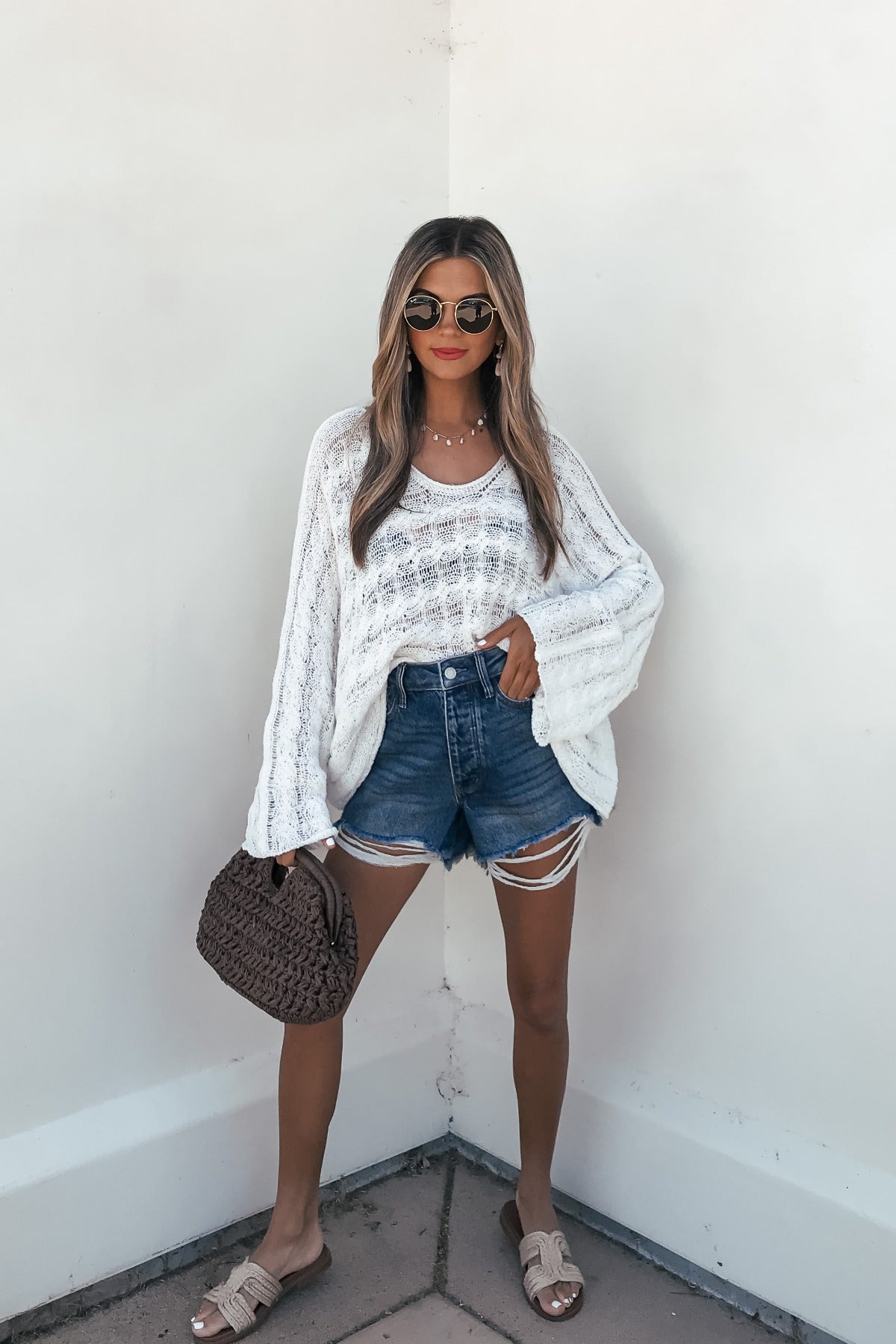 Woman in sunglasses, Calista Open Knit Top - Ivory, denim shorts, and sandals holds a brown bag by a white wall for a breezy coastal look.