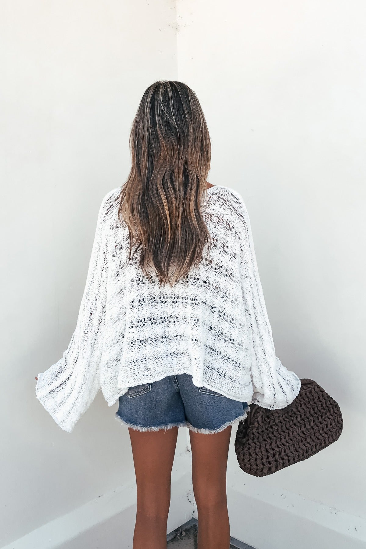 Woman with long wavy hair wears the Calista Open Knit Top in ivory and denim shorts, holding a large brown woven bag by a white wall.