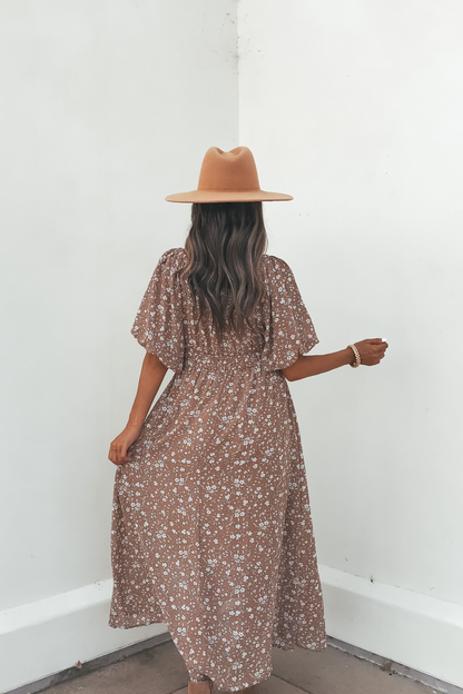 Woman in the Prairie Bloom Maxi Dress - Tan and a tan hat stands by a white wall, holding her dress with one hand.
