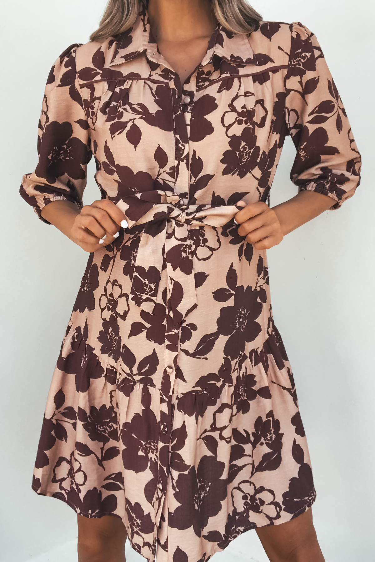 Woman wearing the Lydia Brown Print Tie Front Mini Dress - DOORBUSTER, featuring dark florals and a waist tie, against a white background.