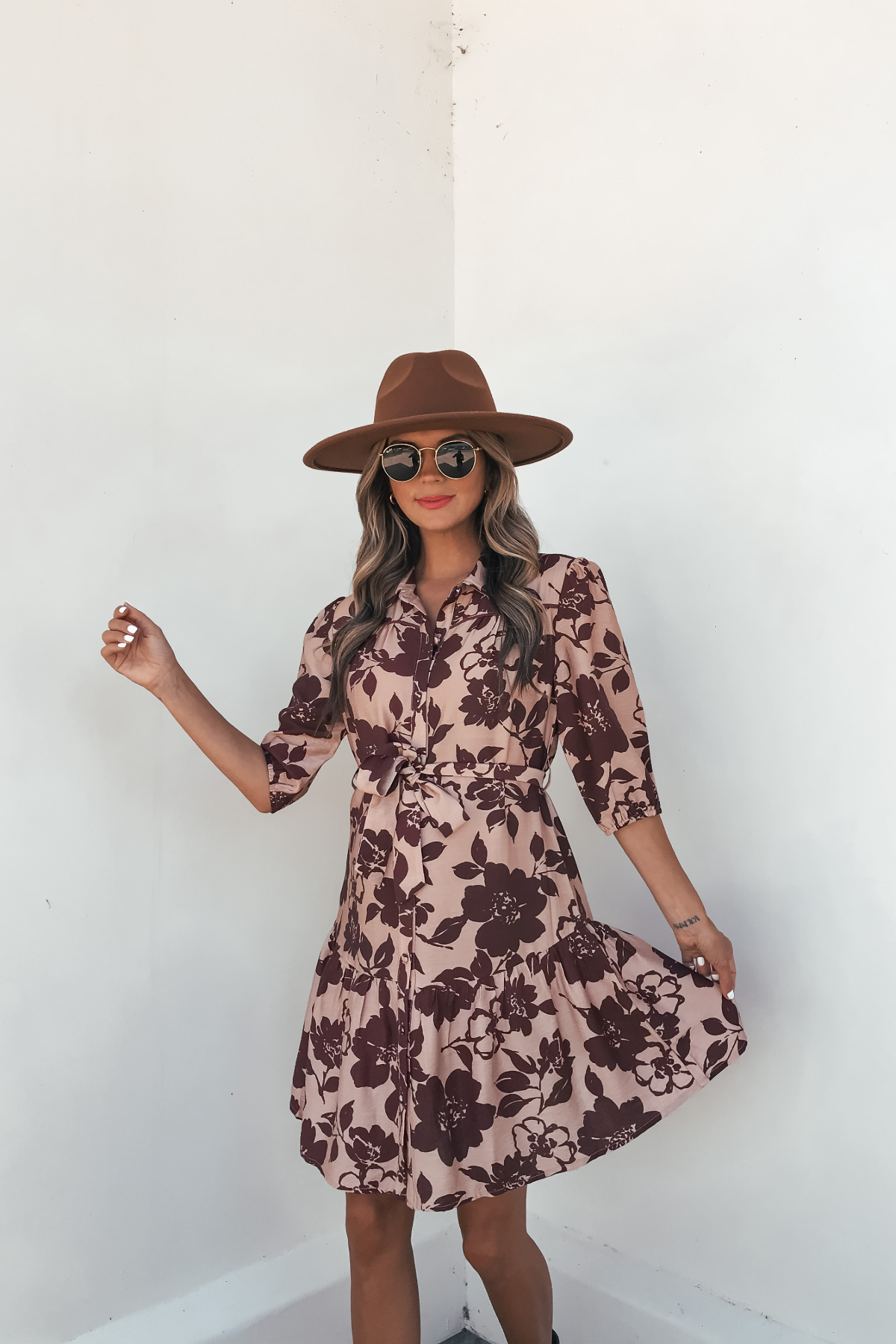 Woman wears the Lydia Brown Print Tie Front Mini Dress - DOORBUSTER, posing against a white wall and holding out the dress.