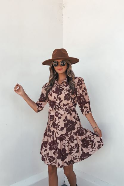 Woman wears the Lydia Brown Print Tie Front Mini Dress - DOORBUSTER, posing against a white wall and holding out the dress.