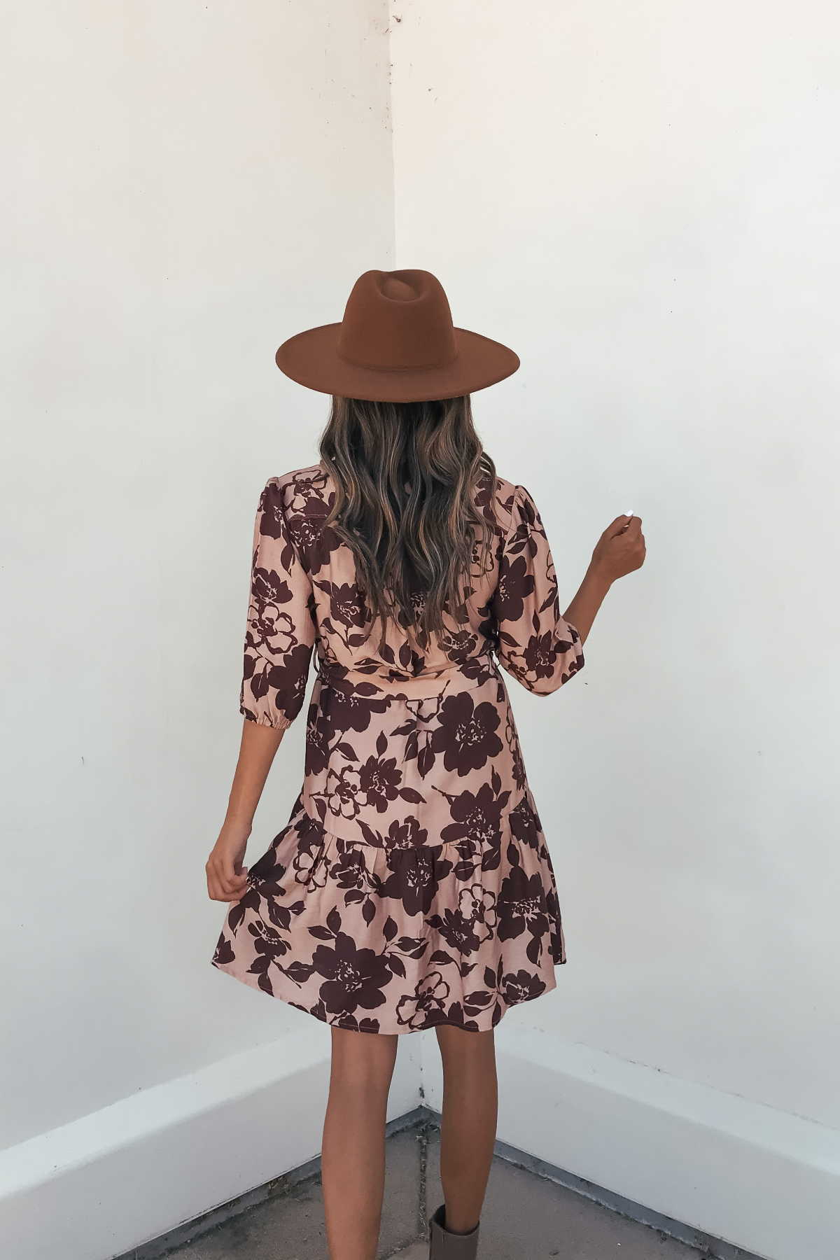 A woman with wavy hair in a brown hat and Lydia Brown Print Tie Front Mini Dress stands with her back to the camera.