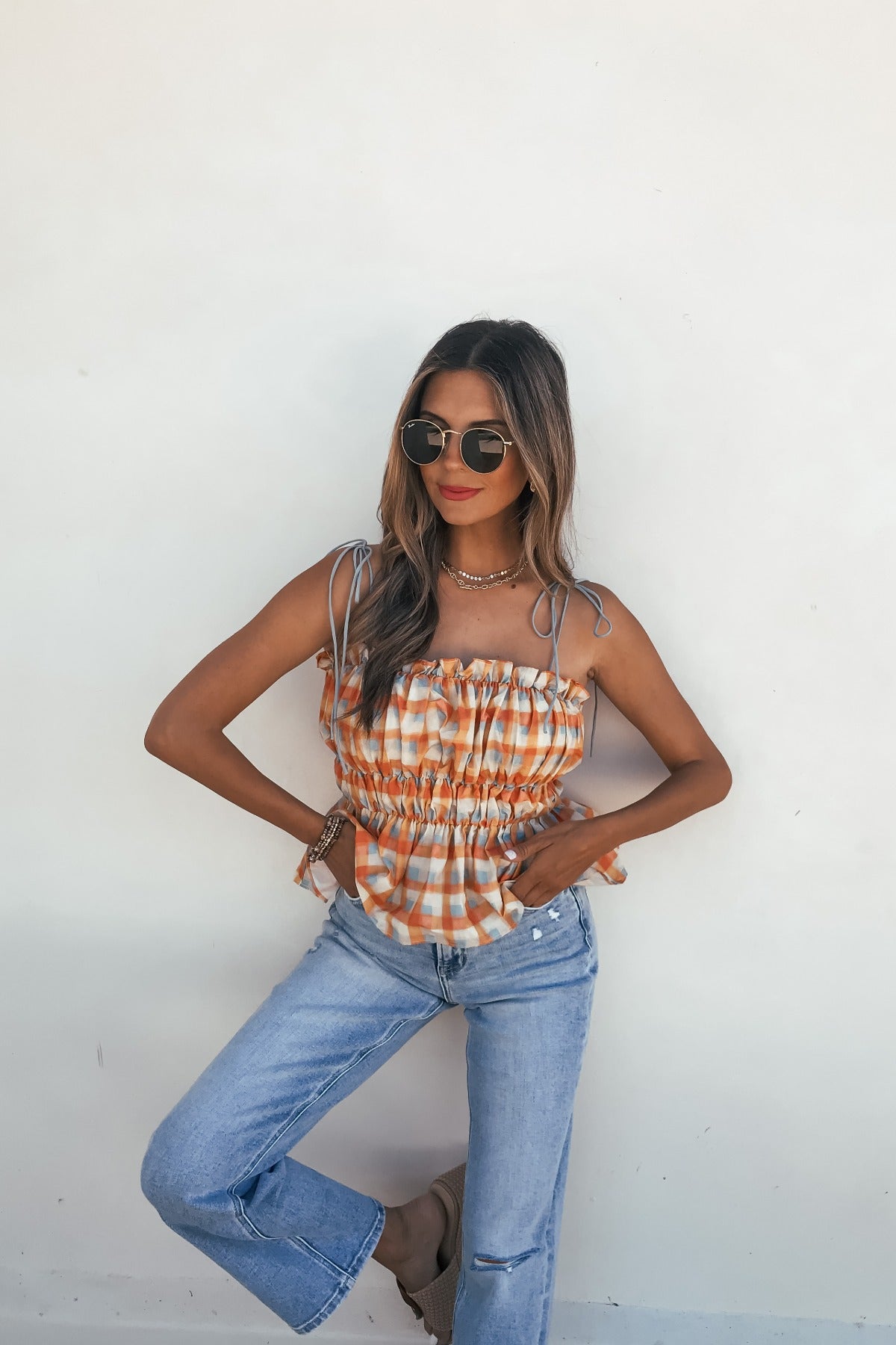Woman in sunglasses wears the Aureum Multi Plaid Smock Top, blue jeans, and brown sandals, posing against a white wall.