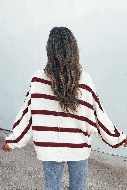 Person with long wavy hair wears a Burgundy Striped Dolman Sleeve Sweater and blue jeans, facing a plain white wall.