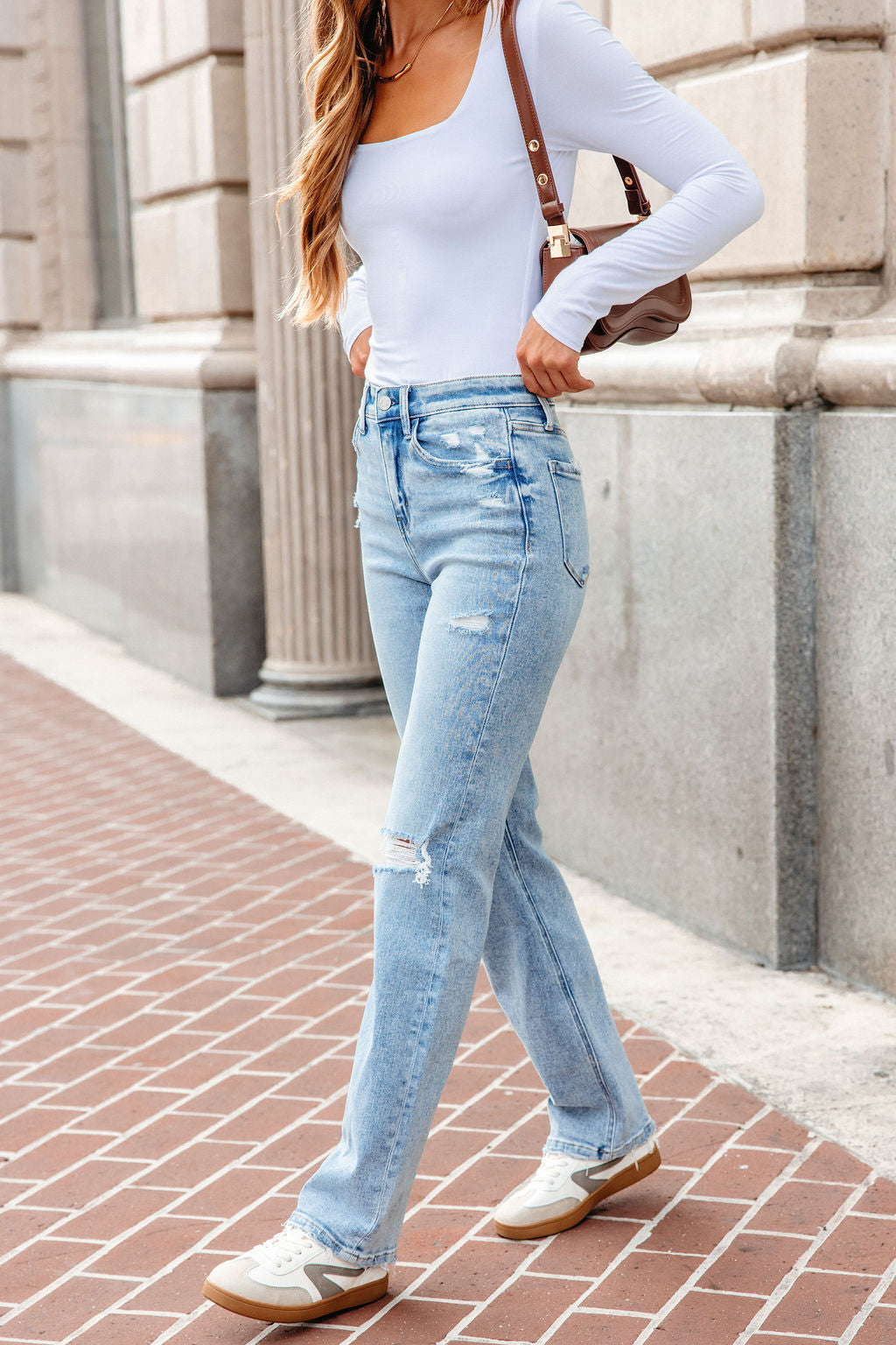 Woman in a white top, Vervet Distressed Straight Leg Jeans - Light Wash, and sneakers walks on a brick sidewalk carrying a brown bag.