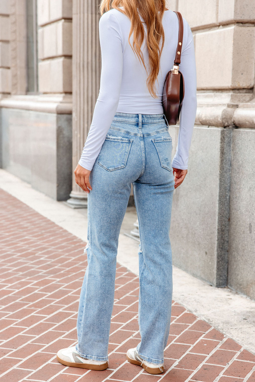Woman in a white long-sleeve top, Vervet Distressed Straight Leg Jeans - Light Wash, sneakers, and a brown shoulder bag on a brick sidewalk.