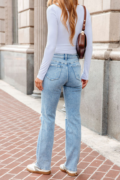 Woman in a white long-sleeve top, Vervet Distressed Straight Leg Jeans - Light Wash, sneakers, and a brown shoulder bag on a brick sidewalk.