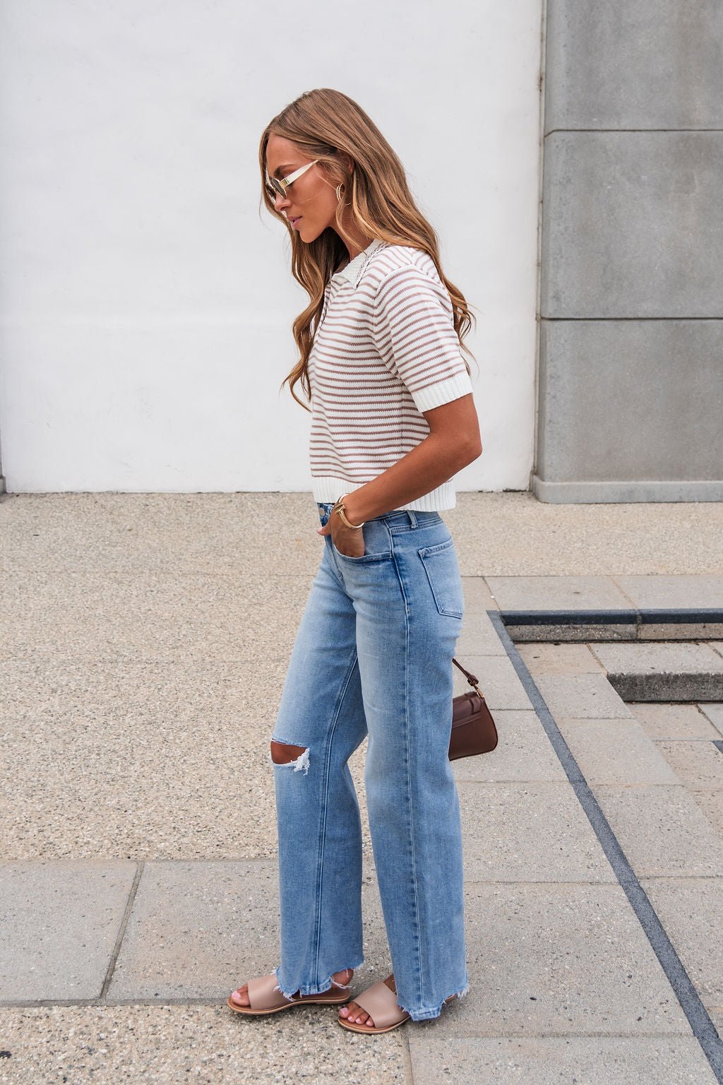 Woman in the Adair Striped Sweater - Dark Taupe, ripped jeans, and sandals stands outside holding a small brown handbag.