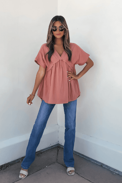 Woman in sunglasses wears the Addison Coral Pleated Tunic Top, blue jeans, and white sandals, standing confidently against a white wall.