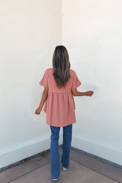 A woman in the Addison Coral Pleated Tunic Top and blue jeans stands with her back to the camera, highlighting this wardrobe essential.