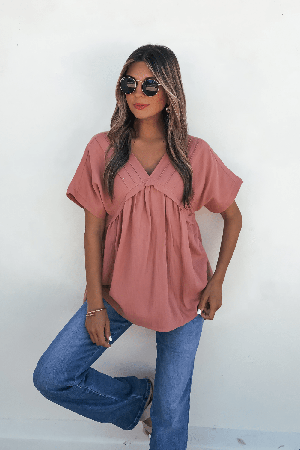 Woman models the Addison Coral Pleated Tunic Top - DOORBUSTER with blue jeans and sunglasses against a plain white background.