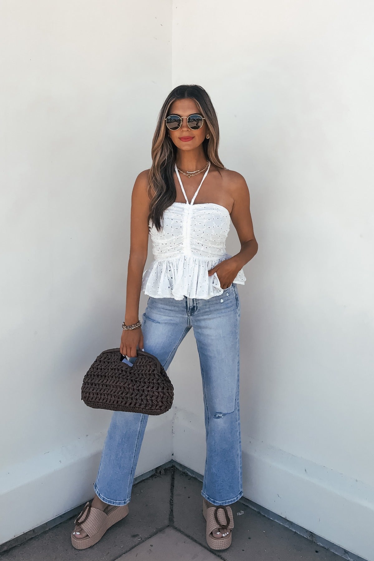 Woman wearing sunglasses, the Adeline Eyelet Smocked Halter Top - White, light blue jeans, and a woven bag—perfect summer style.