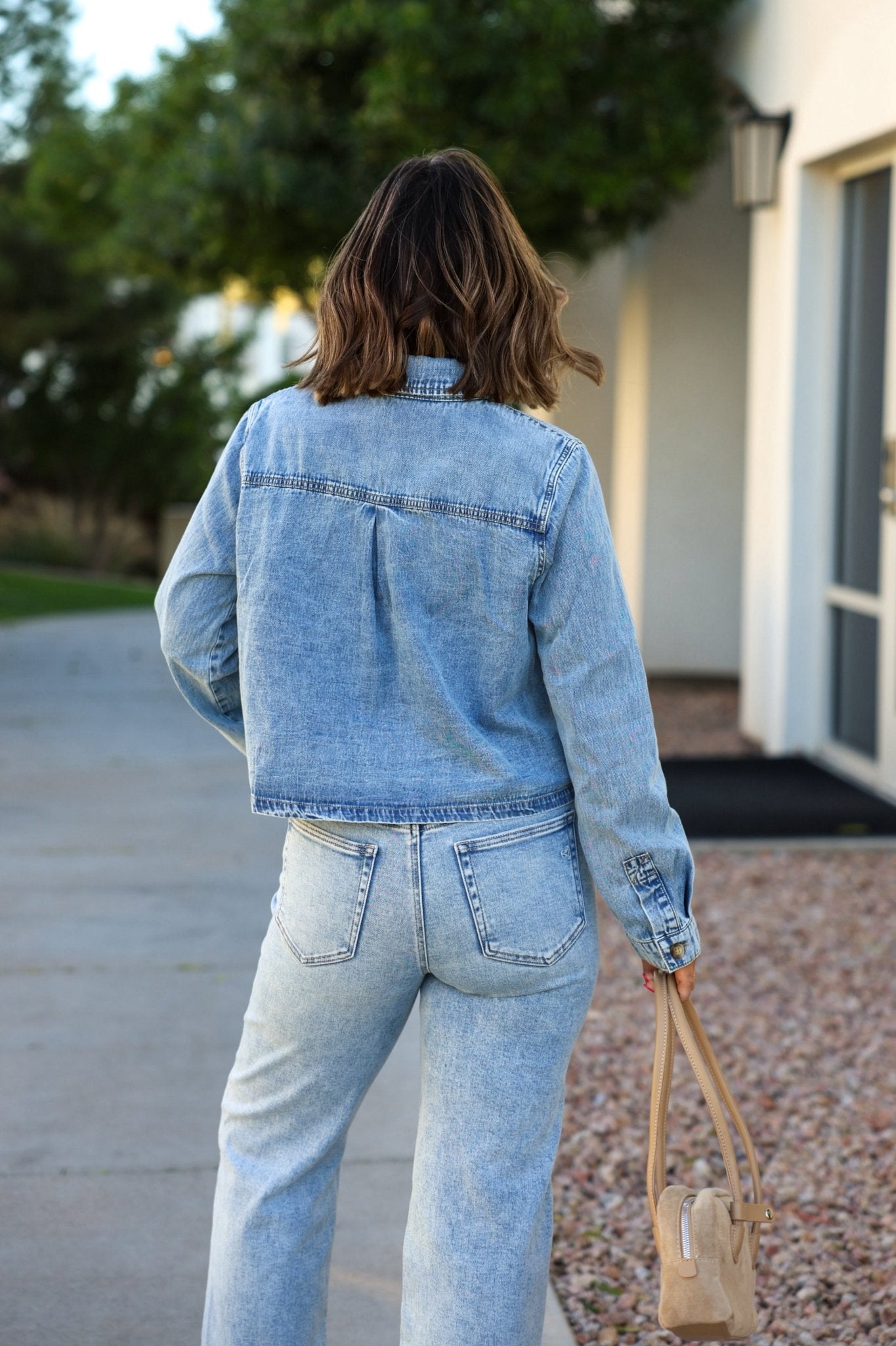 A woman wearing the Adria Boxy Denim Shirt and jeans stands outdoors, facing away, holding a beige handbag in her left hand.