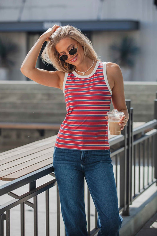 Woman in sunglasses and jeans wears All-Day Stripe Tank Top - Red, holding iced coffee by a railing outdoors.