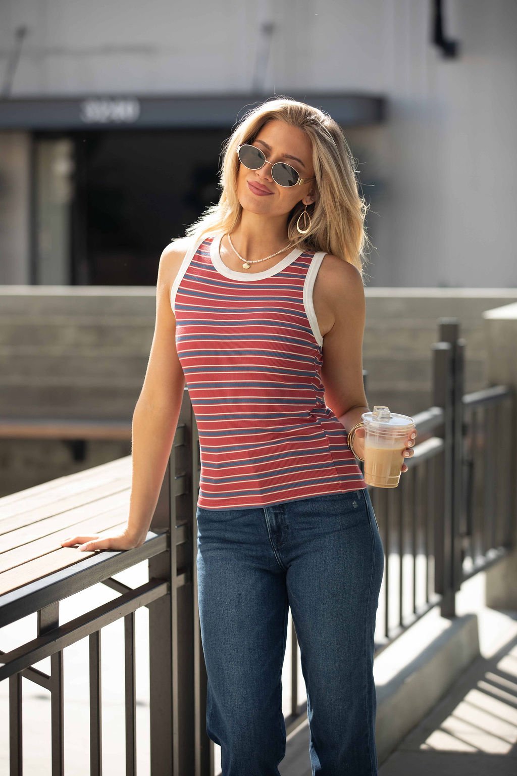Woman in sunglasses and jeans wears the All-Day Stripe Tank Top - Red, holding iced coffee while leaning on a railing outdoors.