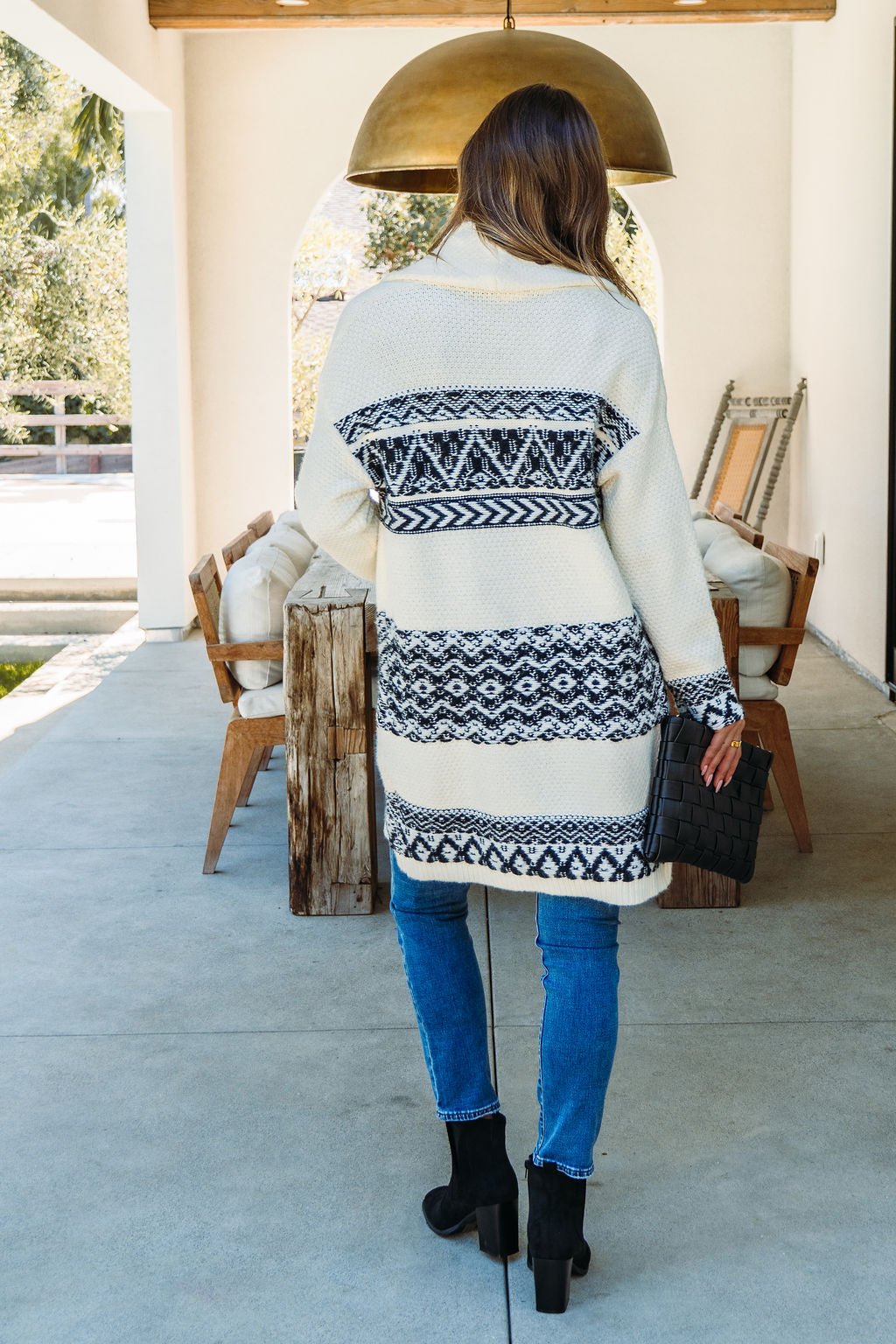 A woman with long hair wears the Alpine Retreat Ivory and Black Shawl Cardigan, jeans, boots, and holds a black handbag on a patio.