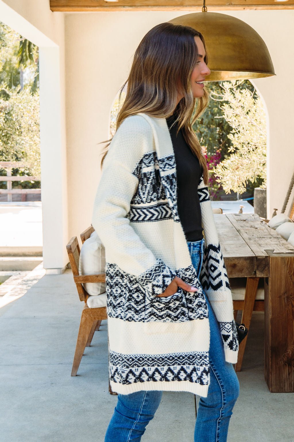 A woman in the Alpine Retreat Ivory and Black Shawl Cardigan stands by a wooden table on a patio, paired with a black top and jeans.
