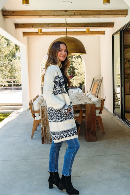 A woman wearing the Alpine Retreat Ivory and Black Shawl Cardigan stands on a covered patio by a wooden dining table.