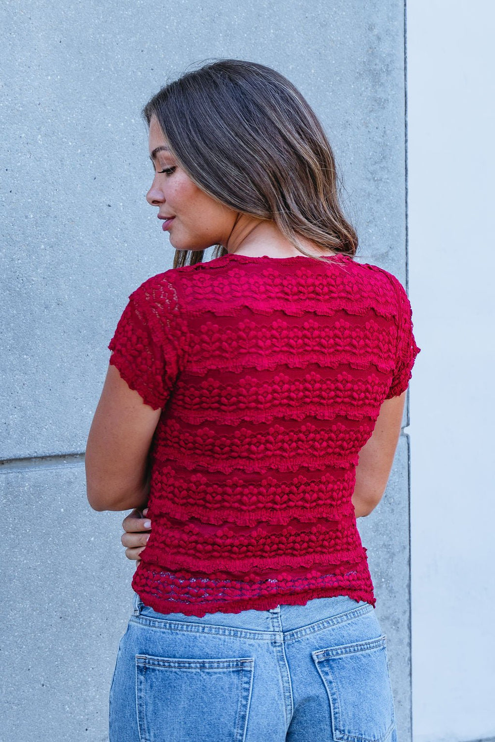 Woman wearing the Amara Burgundy High Neck Lace Top and blue jeans against a gray wall—perfect addition to your fall wardrobe.