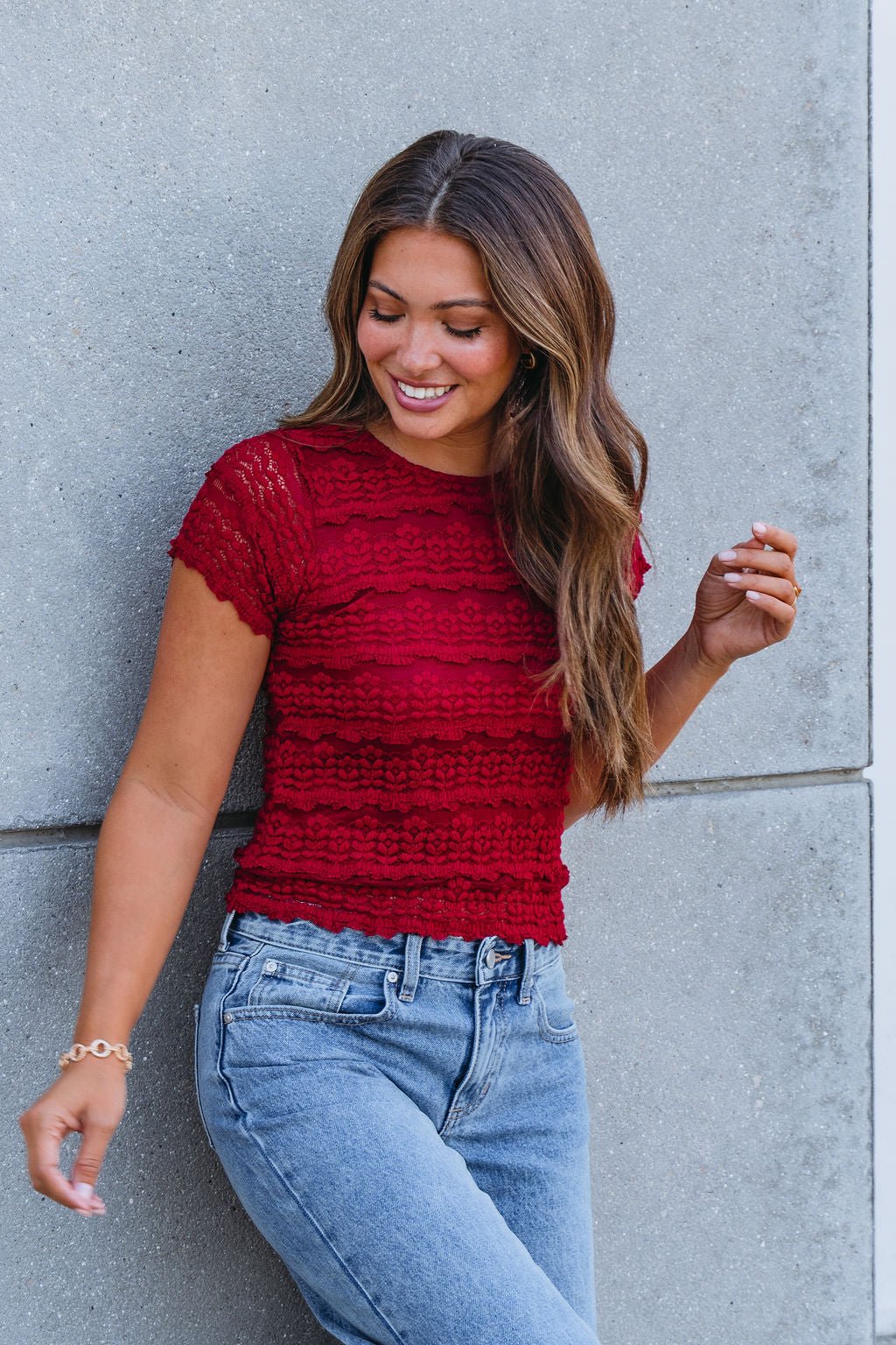 Woman with long brown hair in an Amara Burgundy High Neck Lace Top and blue jeans, smiling by a gray textured wall.