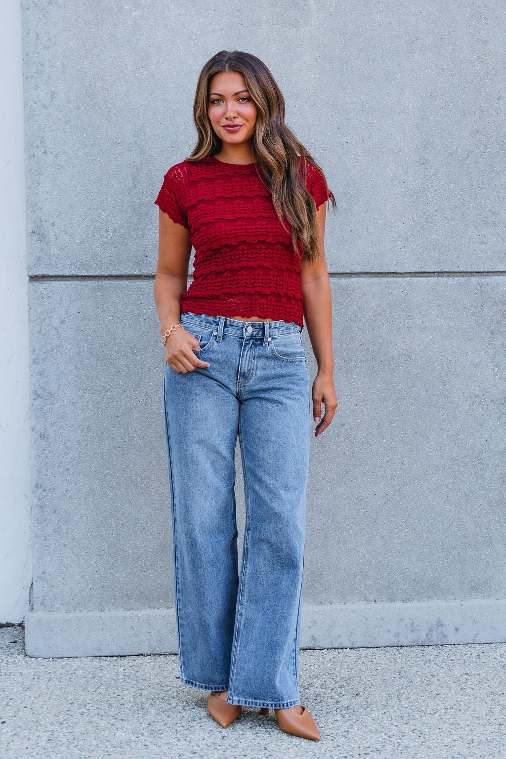 Woman in an Amara Burgundy High Neck Lace Top, blue high-waisted jeans, and tan pointed shoes by a gray concrete wall.