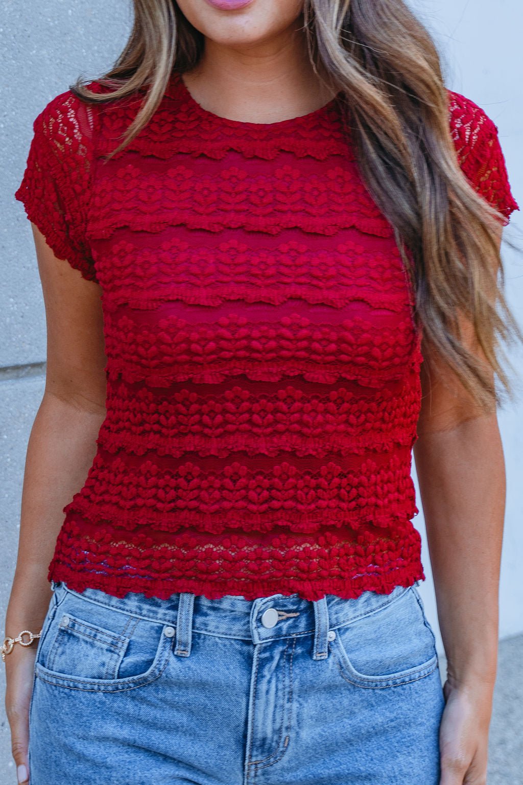 Woman wears the Amara Burgundy High Neck Lace Top with high-waisted blue jeans, perfect for fall, against a light gray wall.